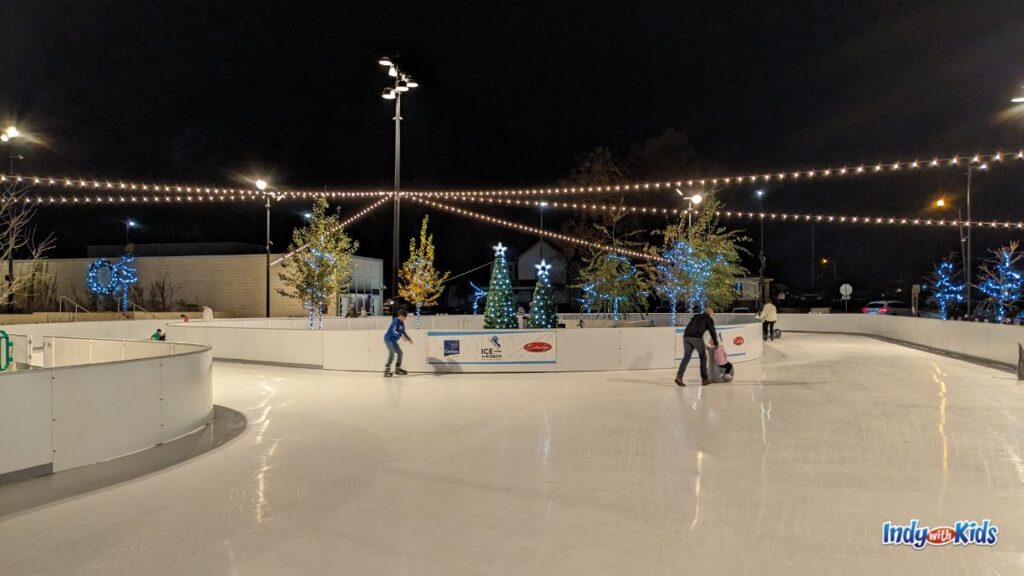 skaters ice skate on the ice ribbon at grand junction plaza Westfield. a dad pushes his child who is sitting on a grey whale ride on. there are string lights coming out from a center pole.