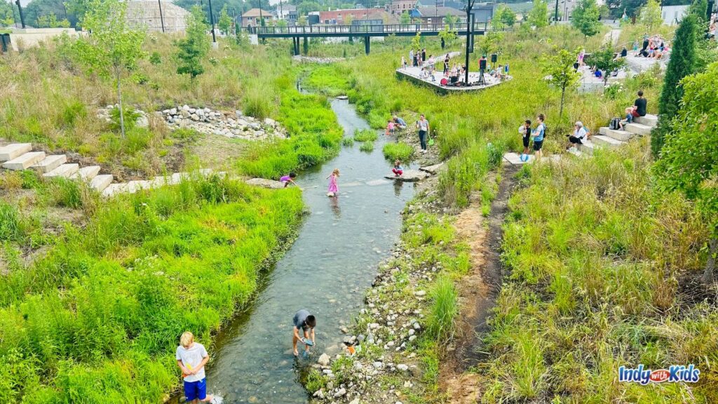 several stone steps lead down to a creek where children are collecting stones and checking out nature at grand junction plaza Westfield. there are plants and tall grass on the banks of the creek and a tall bridge in the background