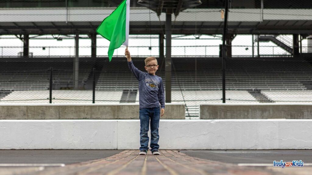 What to Wear to the Indy 500: A boy stands on the yard of bricks at the Indianapolis Motor Speedway. He's wearing jeans and a long sleved shirt while waving a green flag.