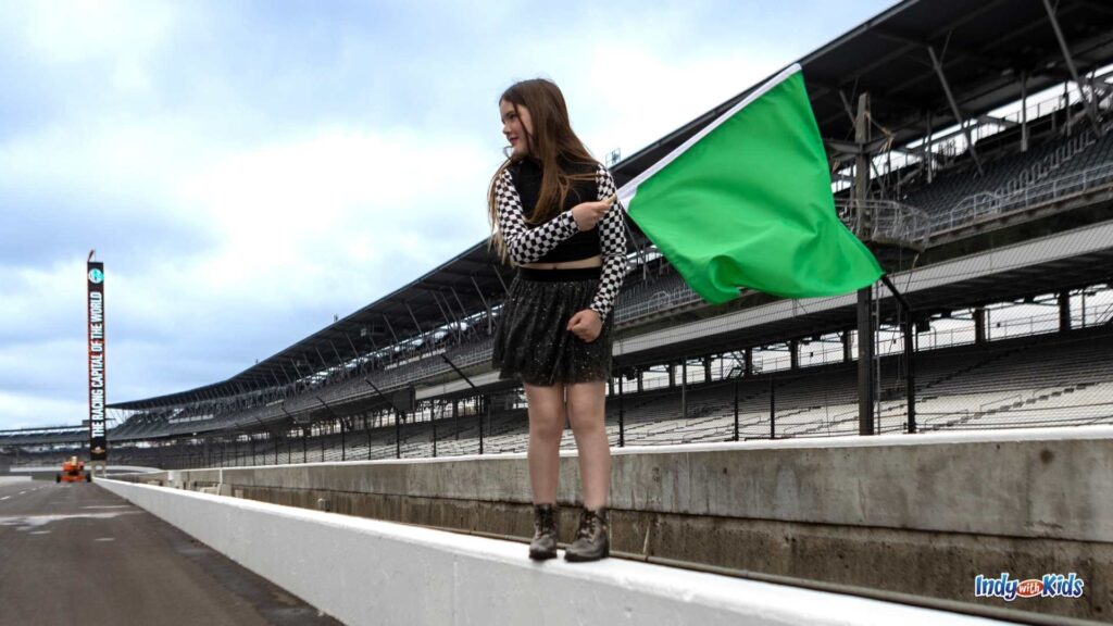 What to Wear to the Indy 500: A pre-teen stands on the wall at the yard of bricks at the Indianapolis Motor Speedway and waves a green flag. She's wearing a black skirt and a black and white checkered crop top.