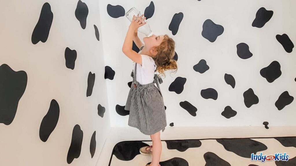 A little girl drinks a giant glass bottle of milk, wearing black and white checked dress, demonstrating what to wear to the Indy 500.