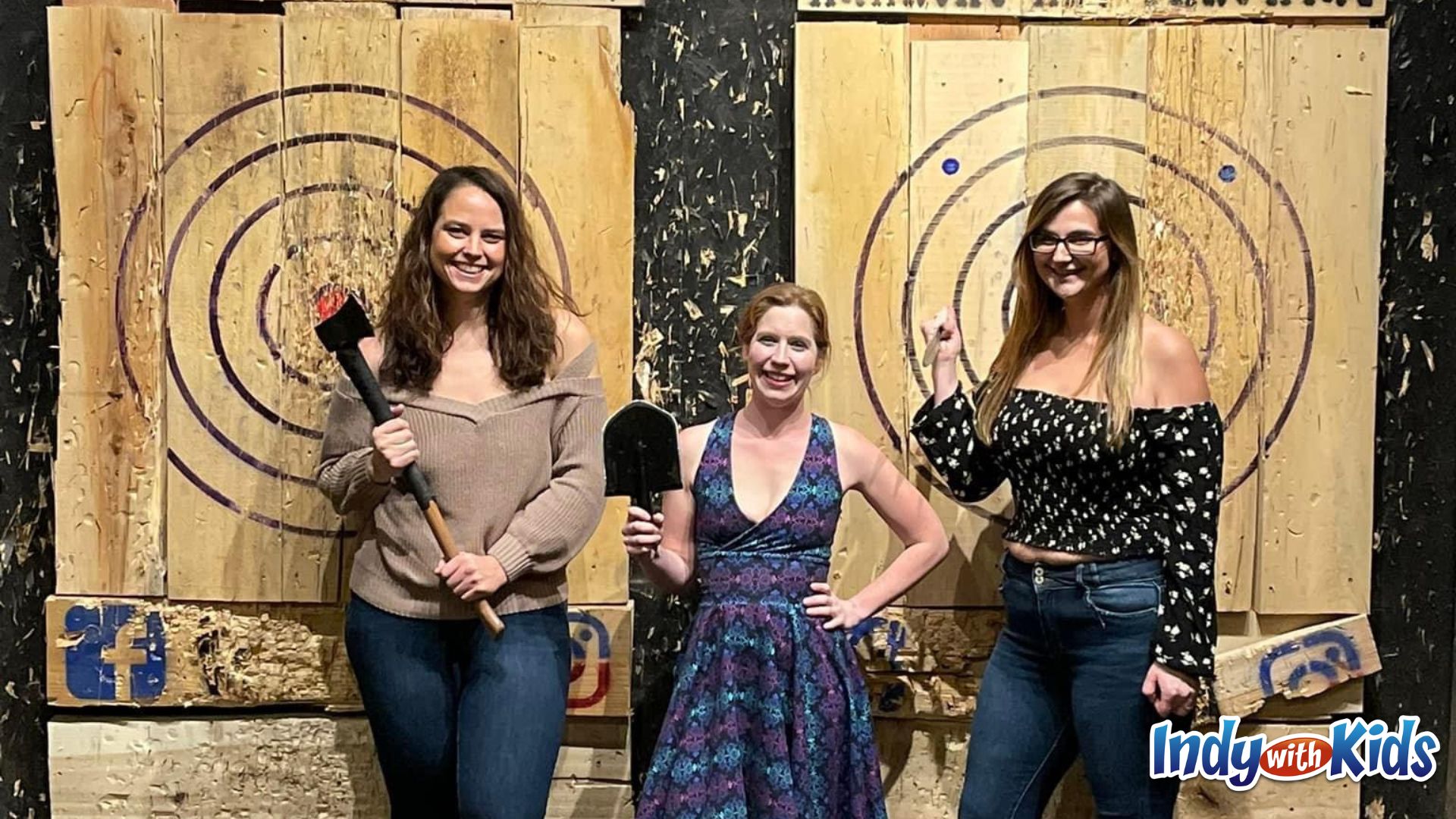 birthday activities for adults: 3 women stand in front of the targets at anarchy axe throwing. one holds an axe, one a shovel, and one a knife.