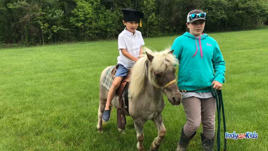 A child wearing a graduation cap rides a small pony while an adult leads the pony through a grassy field.