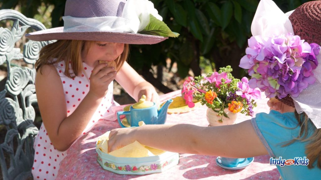 Two young children sit at an outdoor tea party table with hats, flowers, tea cups, and snacks.
