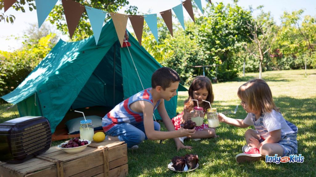Children sit outside near a small tent with picnic snacks, drinks, and party bunting in the yard.