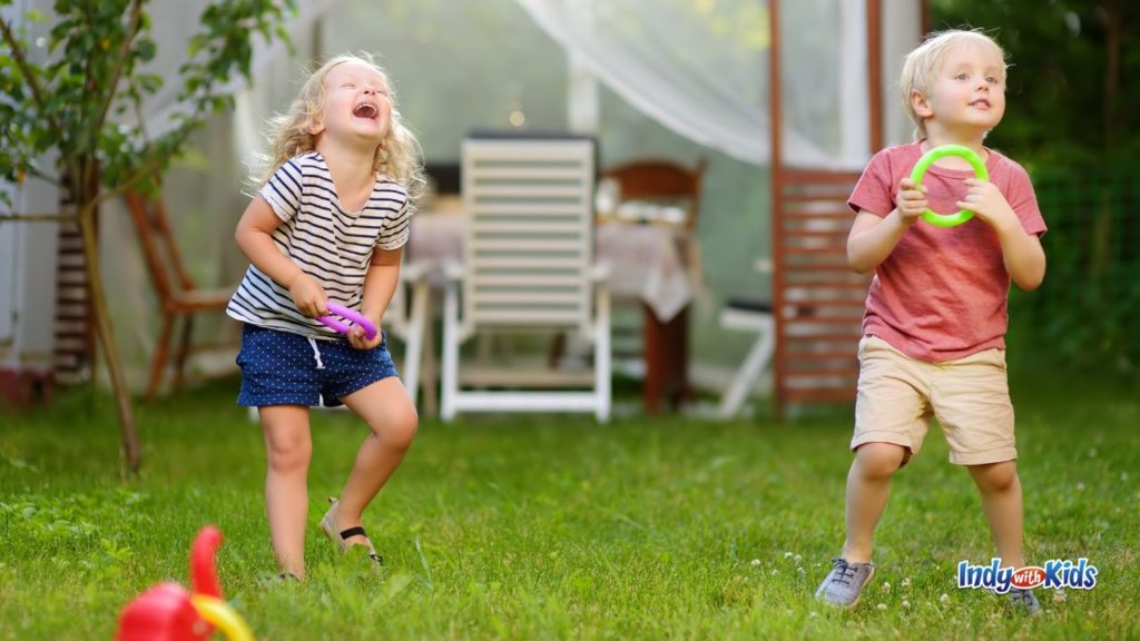 Two children play a ring toss game in a grassy backyard near outdoor patio furniture.