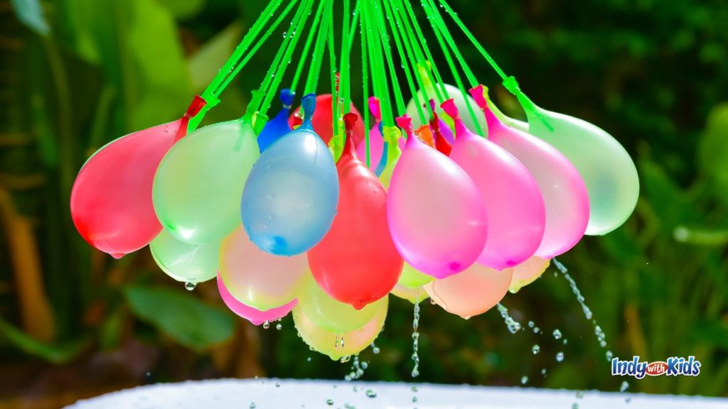 A bunch of colorful filled water balloons hangs together outdoors with water dripping from the balloons.