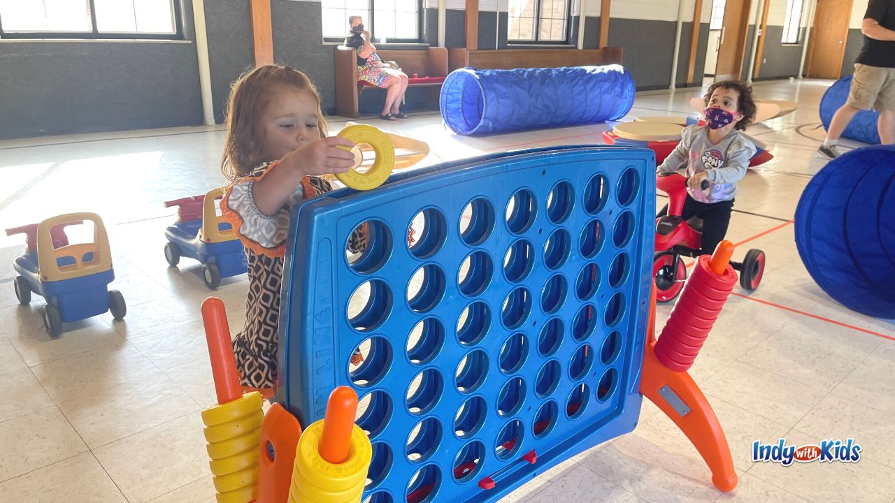 A toddler with red hair plays with a giant connect 4 game set in a gym at Cornerstone Lutheran Church Indianapolis.