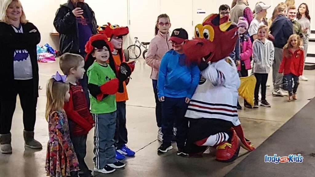the indy fuel mascot, a red dragon, bends down on one knee to greet a line of children, some wearing hats of the mascot