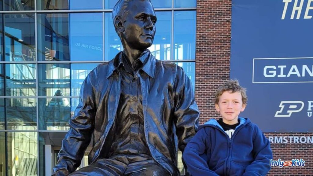 a boy sits next to a bronze statue of a man outside a purdue school building at purdue spring fest