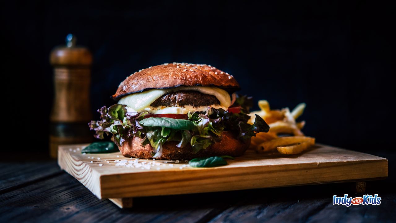The Best Burger in Indianapolis: A hamburger, loaded with greens, cheese, and veggies, sits on a wooden serving board alongside a pile of crispy French fries.