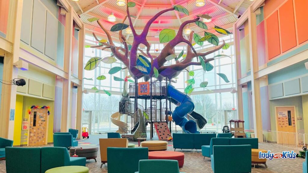 a giant indoor treehouse play structure is in front of a wall of windows in a large indoor playground with colorful chairs and walls
