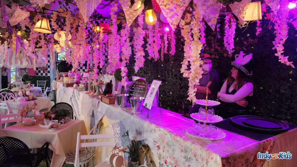 A small room is decorated like a cafe in a faux garden in an Alice in Wonderland themed pop-up bar. Two Alice in Wonderland characters stand behind the bar.