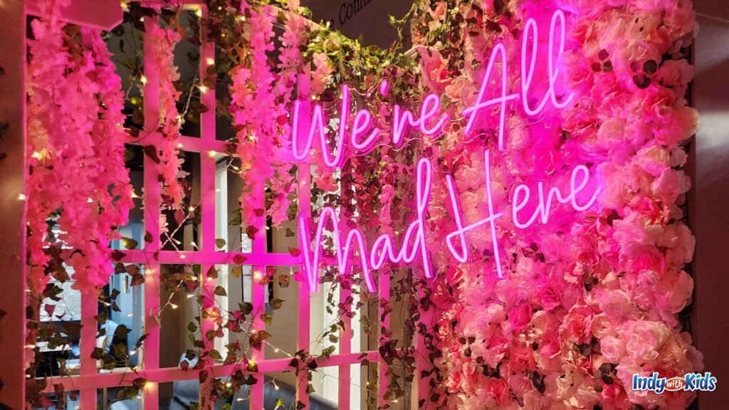 A neon sign glows with pink words that spell out "We're all mad here" on a trellis in a faux garden in an Alice in Wonderland themed pop-up bar.