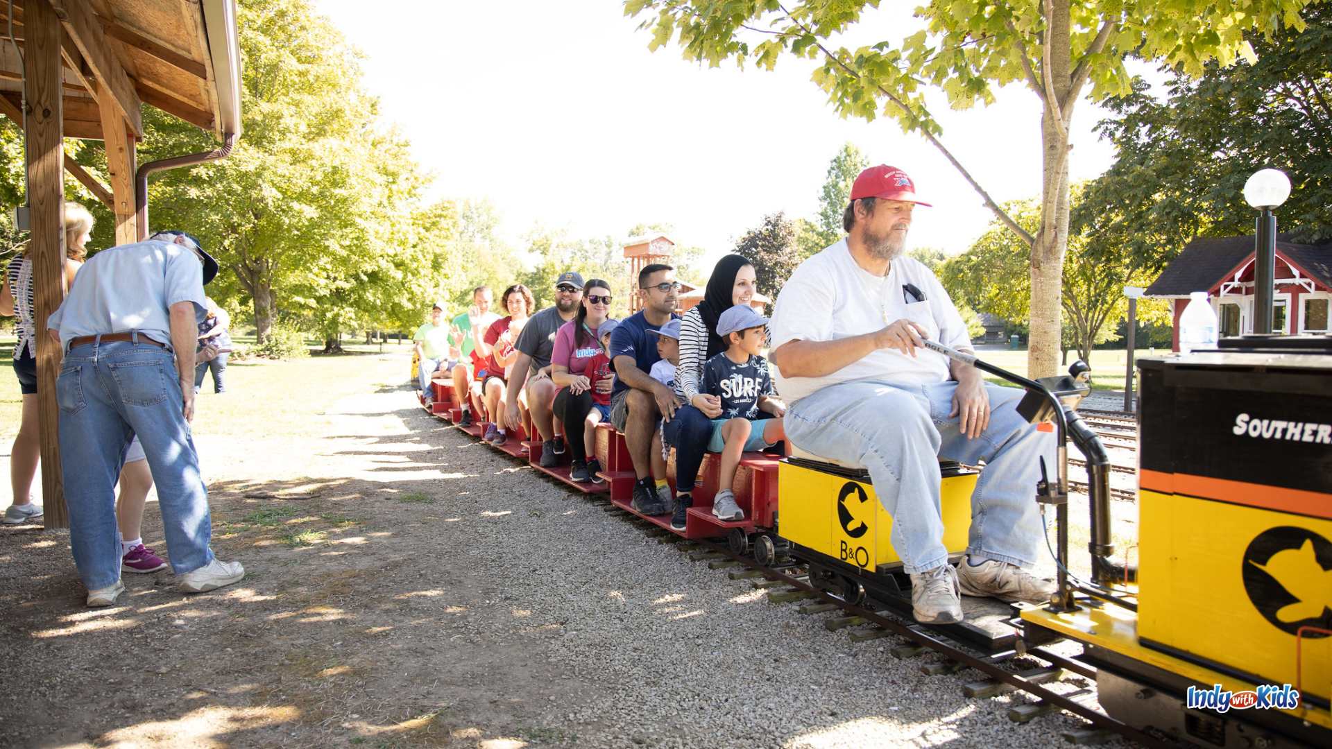 Families ride a working miniature train through a sunny park in Indiana, seated on small open-air train cars behind a yellow and black engine.