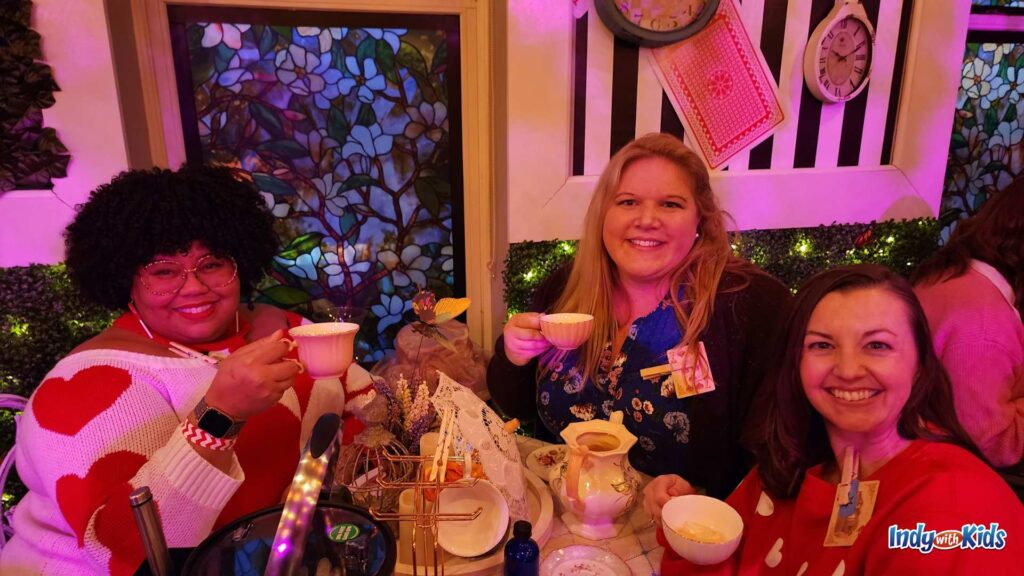 Three women holding teacups up at a cafe table, surrounded by a faux garden in a Alice in Wonderland themed pop-up bar.
