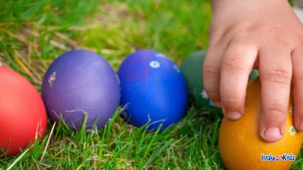 A toddler's hand reaches for an orange dyed egg in the green grass in our list of Easter Egg Hunts in Indiana.