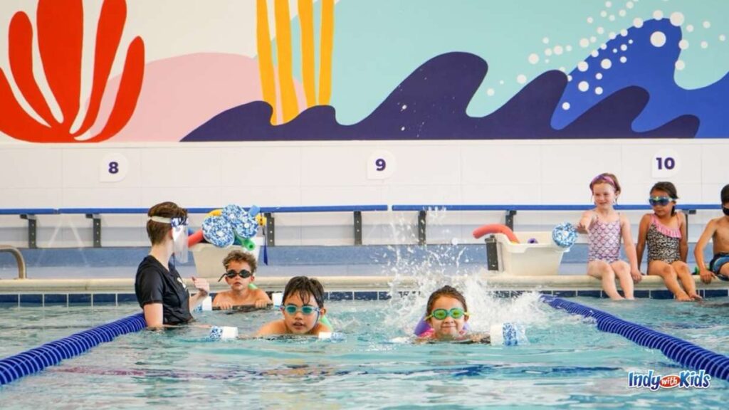 Two children with goggles on practice swimming the length of the pool while an instructor hangs back with another child and three children in the next lane look on