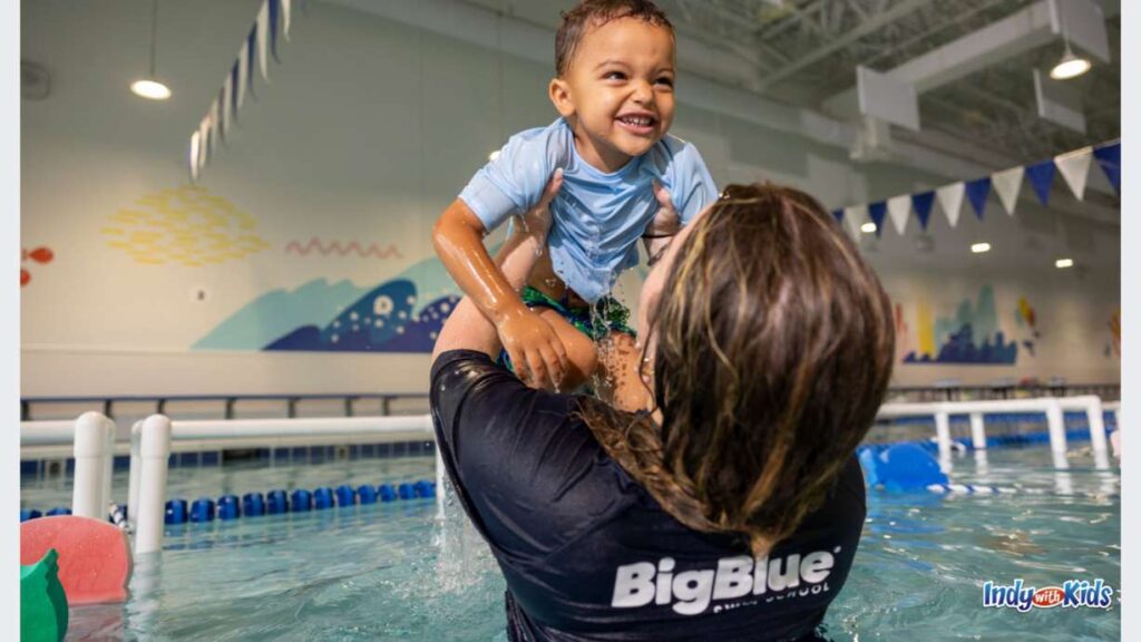 A Big Blue swim instructor holds up a smiling baby boy in the pool during a swim lesson
