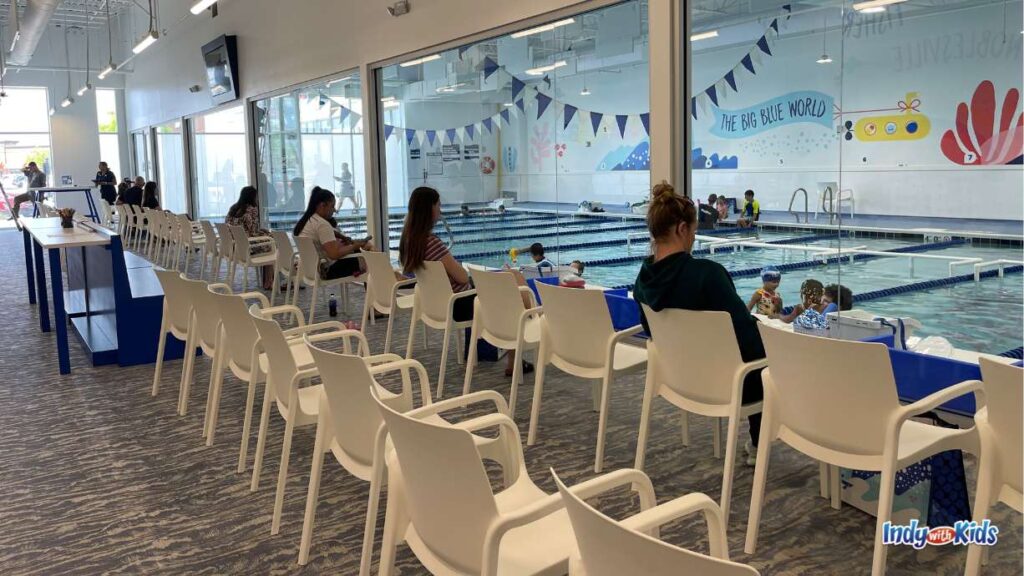 Parents sit on white chairs facing a wall of windows overlooking the Big Blue swimming pool where children are taking swim lessons in lanes divided by blue dividers