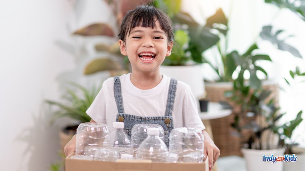 A young girl with bangs and overalls grins happily while holding a cardboard box full of clean plastic bottles, ready to be recycled for Earth Day.