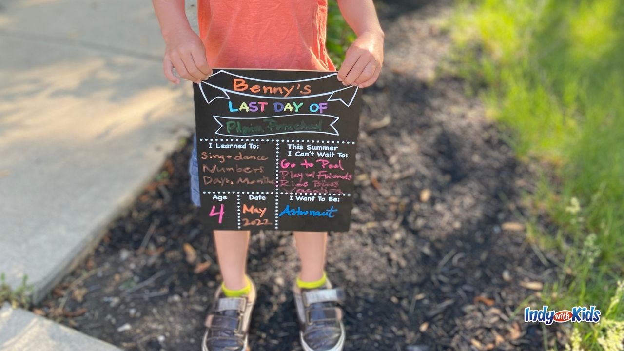 a child wearing a salmon colored shirt and shorts holds up a black chalkboard sign with information about his interests and hopes for the summer