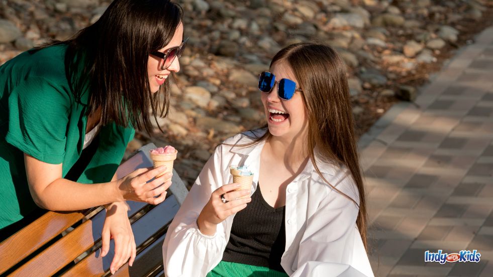 A teen and her mom, wearing sunglasses, laugh and eat ice cream on Mother's Day.