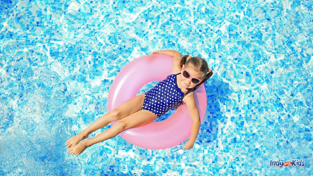 a young girl floats in a pool on a pink donut inner tube. she is wearing a blue and white polka dotted swim suit and pink rimmed sunglasses