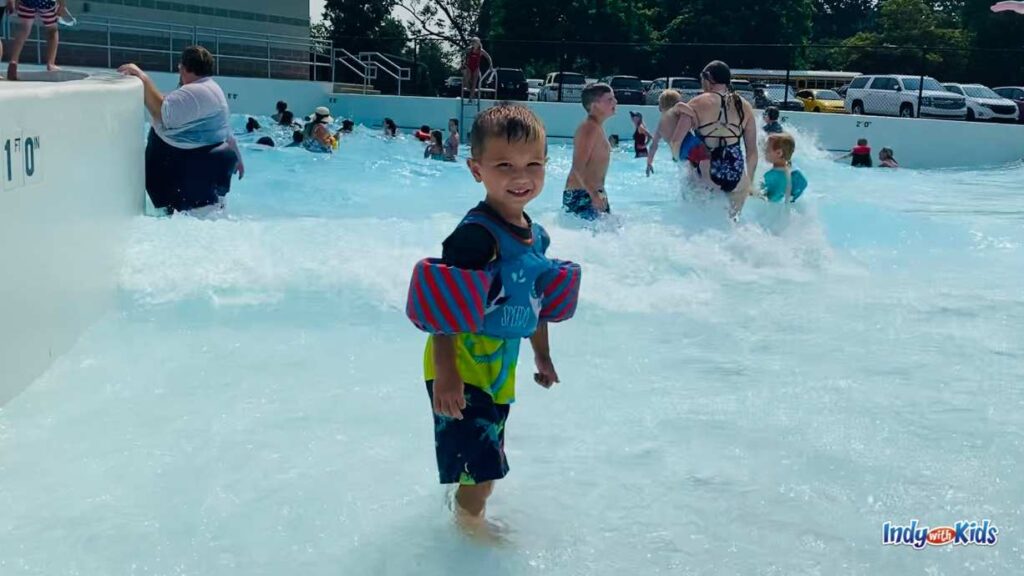 a little boy wearing a puddle jumper smiles as he stands ankle deep in a wave pool. several people behind him are in deeper water in the wave pool.