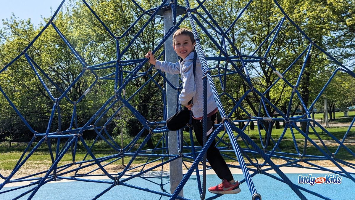 Mud free parks near me: A boy smiles while climbing a blue spider-web rope structure at O'Bannon Park in Indianapolis.