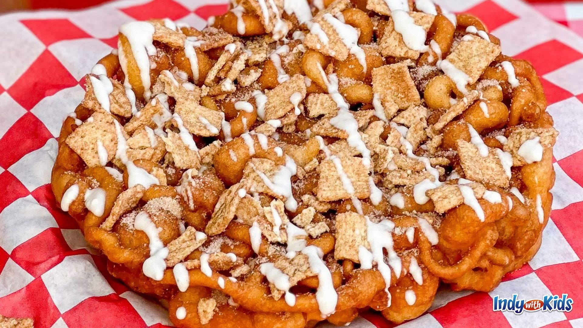 Indiana State Fair Food: A funnel cake on a red and white checkered paper is topped with cinnamon toast crunch cereal and a drizzle of vanilla frosting.