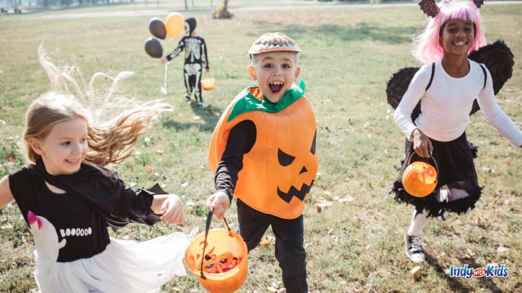 Whitestown Trick or Treat Trail | Halloween 2024: children run through a grass field dressed up as a pumpkin, ghost, witch, and skeleton. all holding pumpkin buckets.