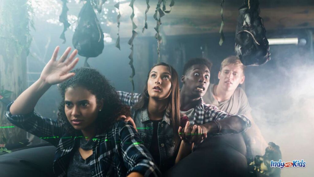 Haunted House Near Me: 4 teens walk through a haunted house with streamers and other object hanging from the ceiling. they all look very scared.