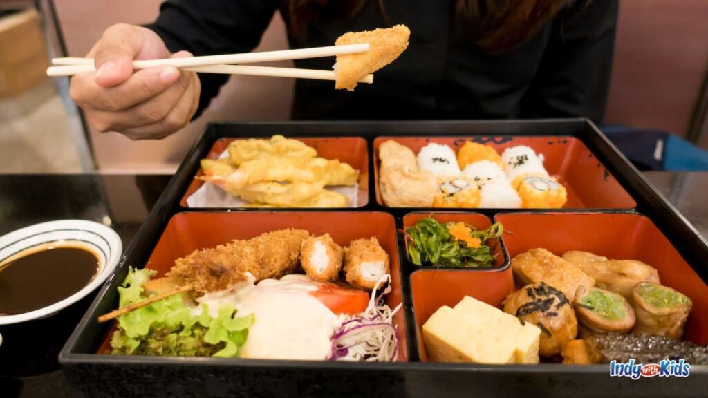 Devour Indy: a person in a black shirt holds a piece of chicken above a red bento box full of Japanese fare with chopsticks