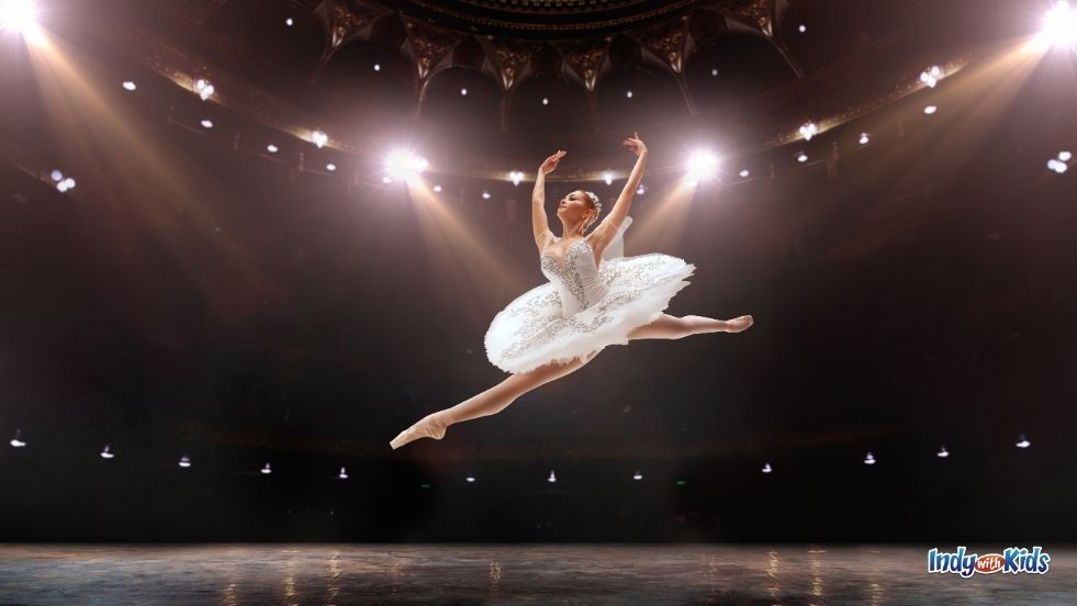 A ballerina in a white tutu leaps across a stage, arms raised, as spotlights illuminate the space.
