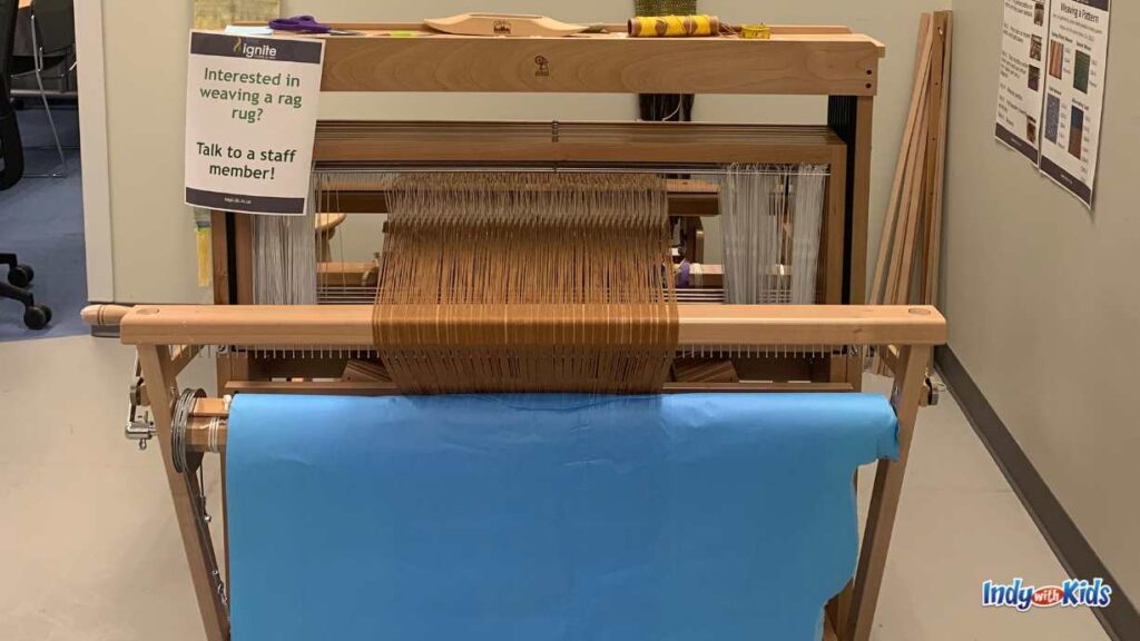a large wooden loom with blue paper hanging down the front sits in front of three finished pieces on the wall. a laminated sign reads "interested in weaving a rag rug? talk to a staff member!"