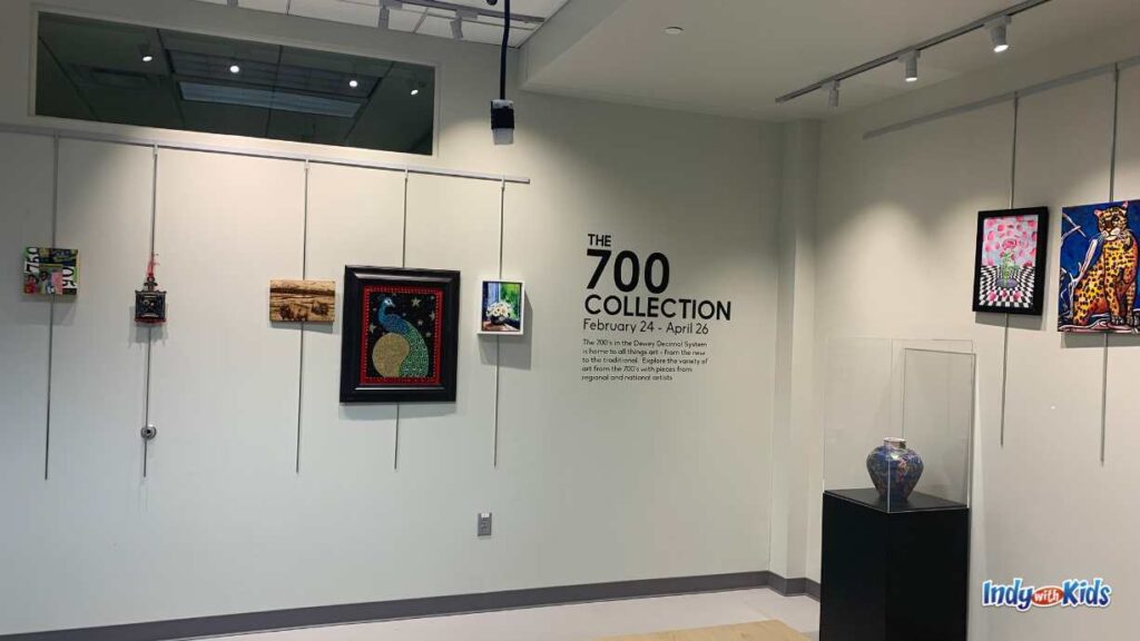 an exhibit titled "the 700 collection" is displayed. the title and information is written on the wall and several pieces of art hang down the walls. there is also a blue vase in a case in the room.