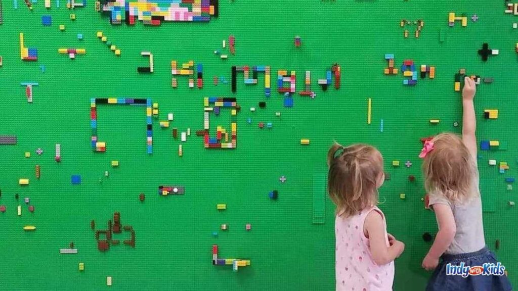 two little girls place legos on a green lego wall full of creations left behind by other children