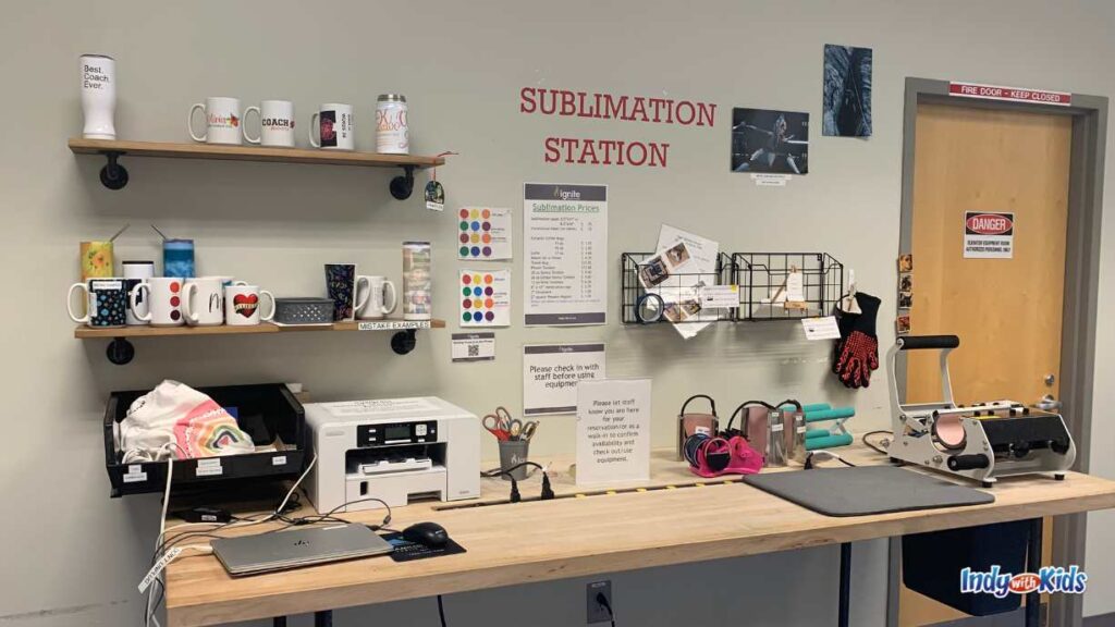 the sublimation station has a wooden table against a wall that holds computers, printers, a clam press, and other equipment. there are wall shelves holding cups, water bottles, and other products to engrave and press.