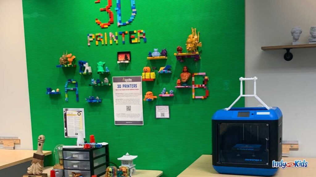 a green wall showcases the words "3D printer" made out of legos. Underneath on the wall are colorful 3D creations. A 3D printer sits on a table next to the wall. 