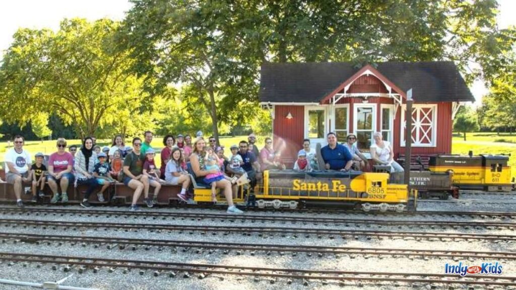 children and adults sit on a mini train on tracks at indiana live steamers