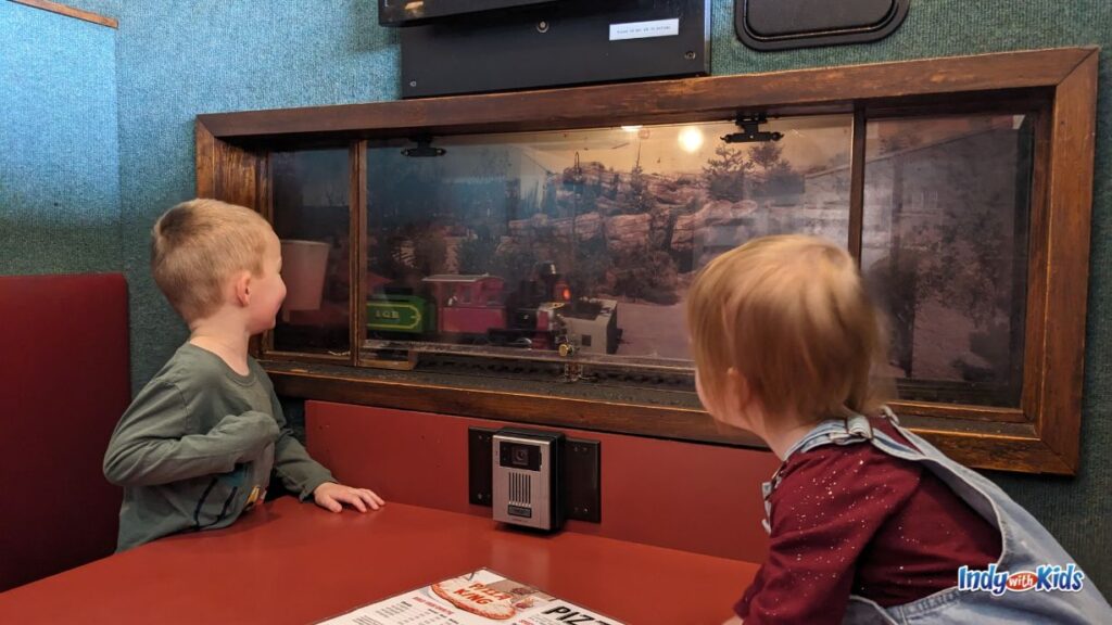 two boys watch through a window as a train delivers their drinks to their table at pizza king