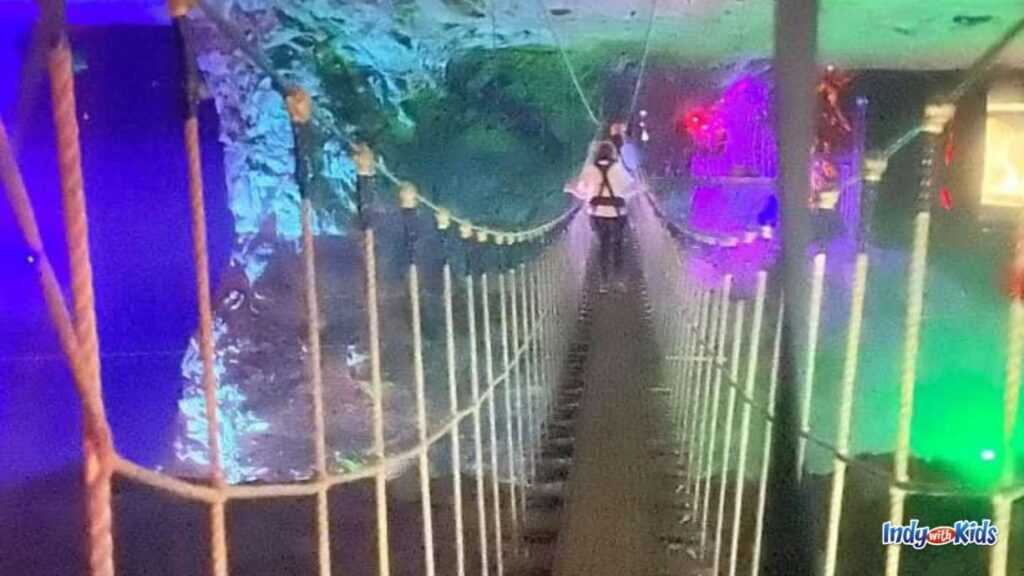 a person wearing a helmet walks across ropes course bridge in the underground Cavern. the Cavern is lit up in blue and green.