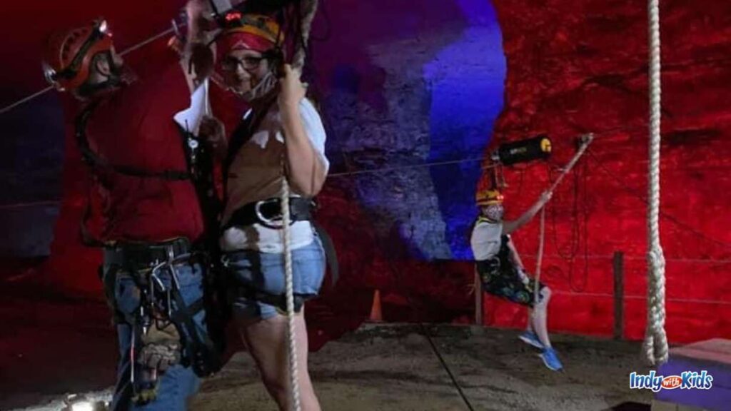 two ladies are getting harnessed for the Zipline tour underground at Louisville Mega Cavern ziplines. they have helmets on and the rock behind them is lit up red.