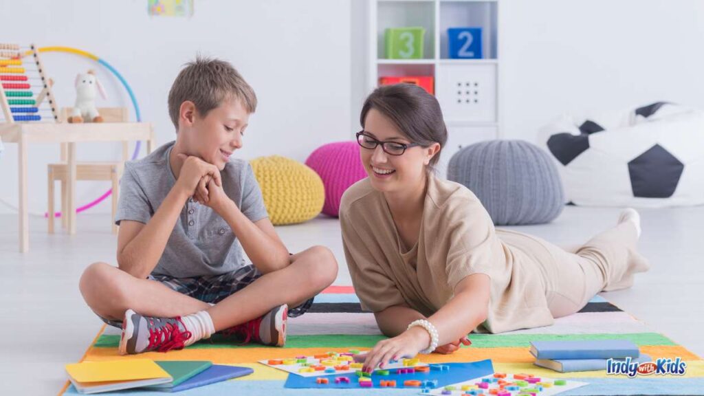 a babysitter lays on the ground in a room with colorful decor pushing around letter magnets next to a boy sitting and smiling. 