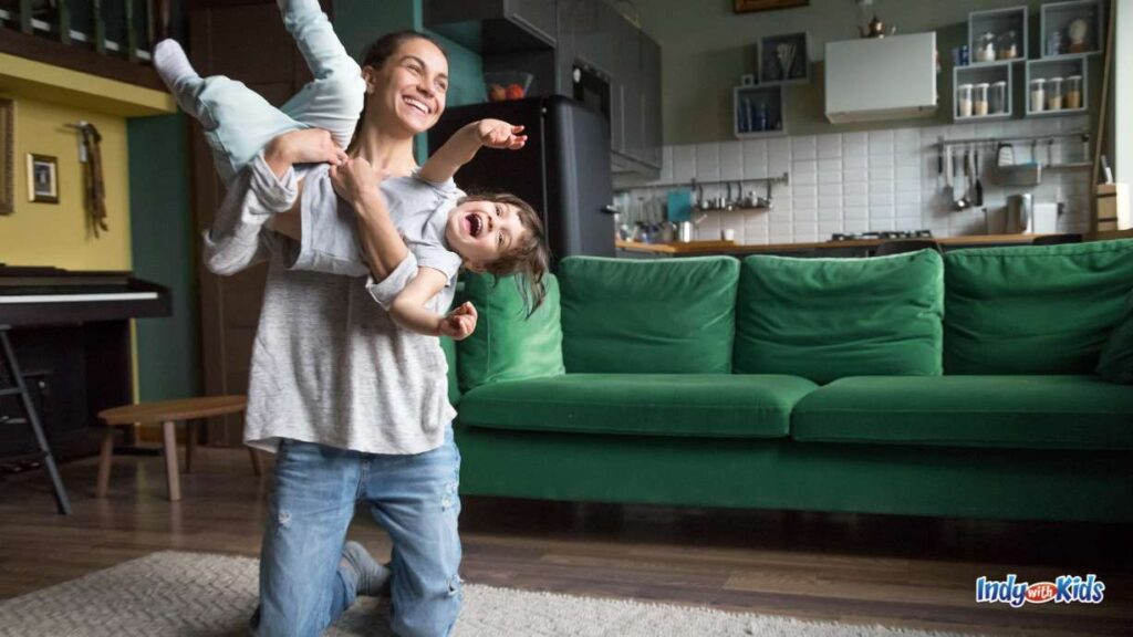 a woman holds up a child (who is horizontal with both legs in the air) with both arms in the living room of a home with a green sofa and the kitchen in the background. both are smiling.