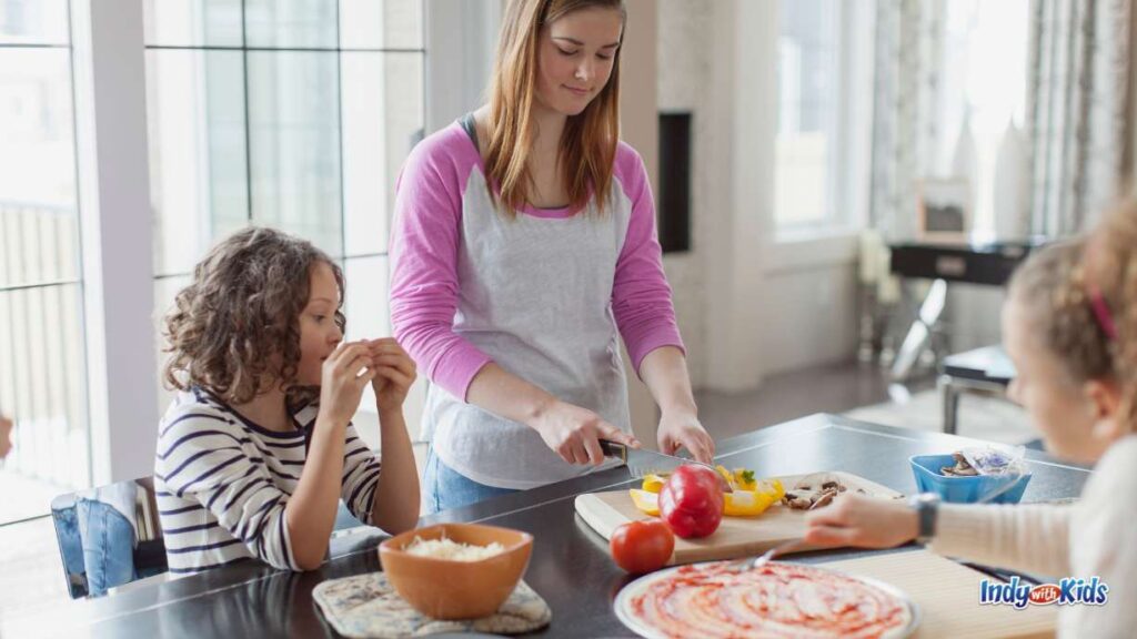 a young lady cuts red and yellow peppers at the kitchen island with two school-aged girls. One girl is spreading marinara sauce on a pizza and one is eating a snack.