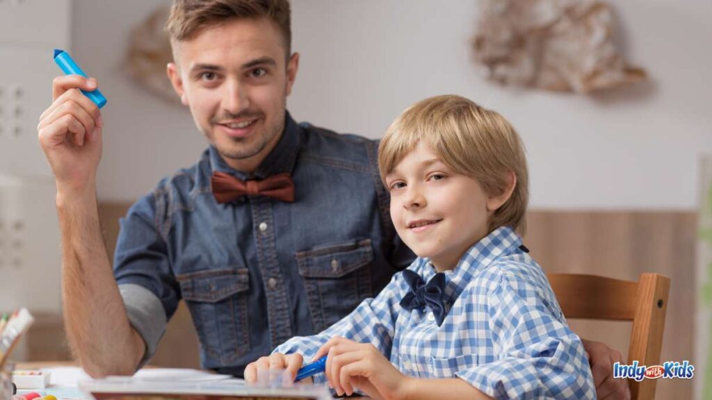 a male (who could be a babysitter or tutor) with a jean shirt and red bowtie, holding a blue marker, sits next to a boy with a blue shirt and blue bowtie at a table doing homework.