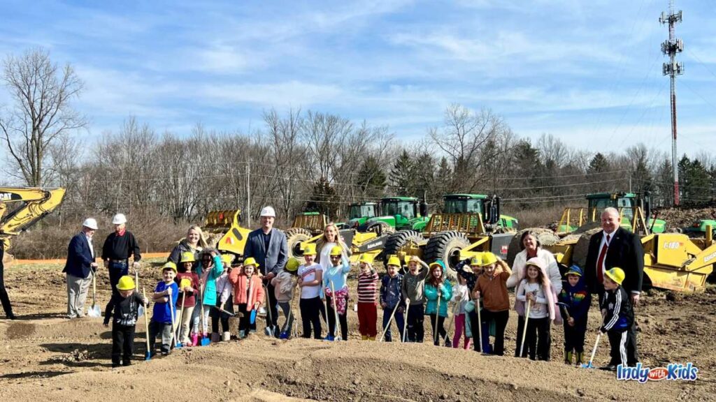 adults and children stand on a dirt plot with shovels in hand and hard hats on with green and yellow construction diggers in the background