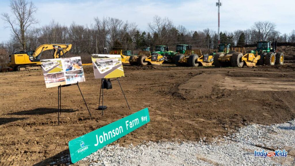 a street sign that says Johnson Farms Way sits on the ground at a construction site in front of two poster renderings on easels and several construction vehicles in the background 